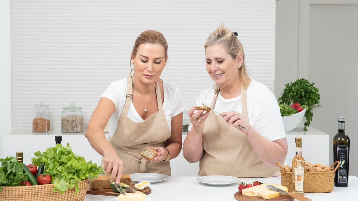 Two women prepare sandwiches with fresh vegetables and bread, enjoying a cooking session together indoors.